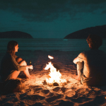 late night beach scene with two people watching water with raging campfire in front shot