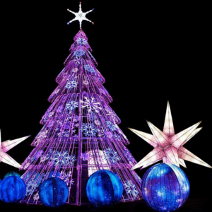 dark night sky with large lit Christmas tree sculpture in purple, blue and white with large blue baubles on floor surrounding. 